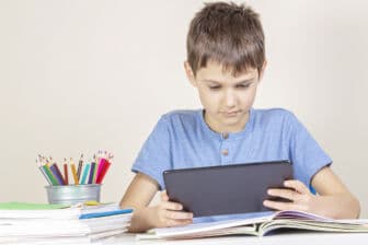 Kid with tablet computer sitting at table with books notebooks.