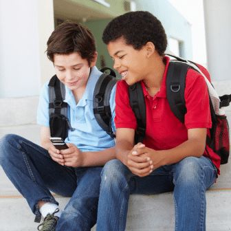 Two teens with backpacks on sit on steps, one looking at the other's phone